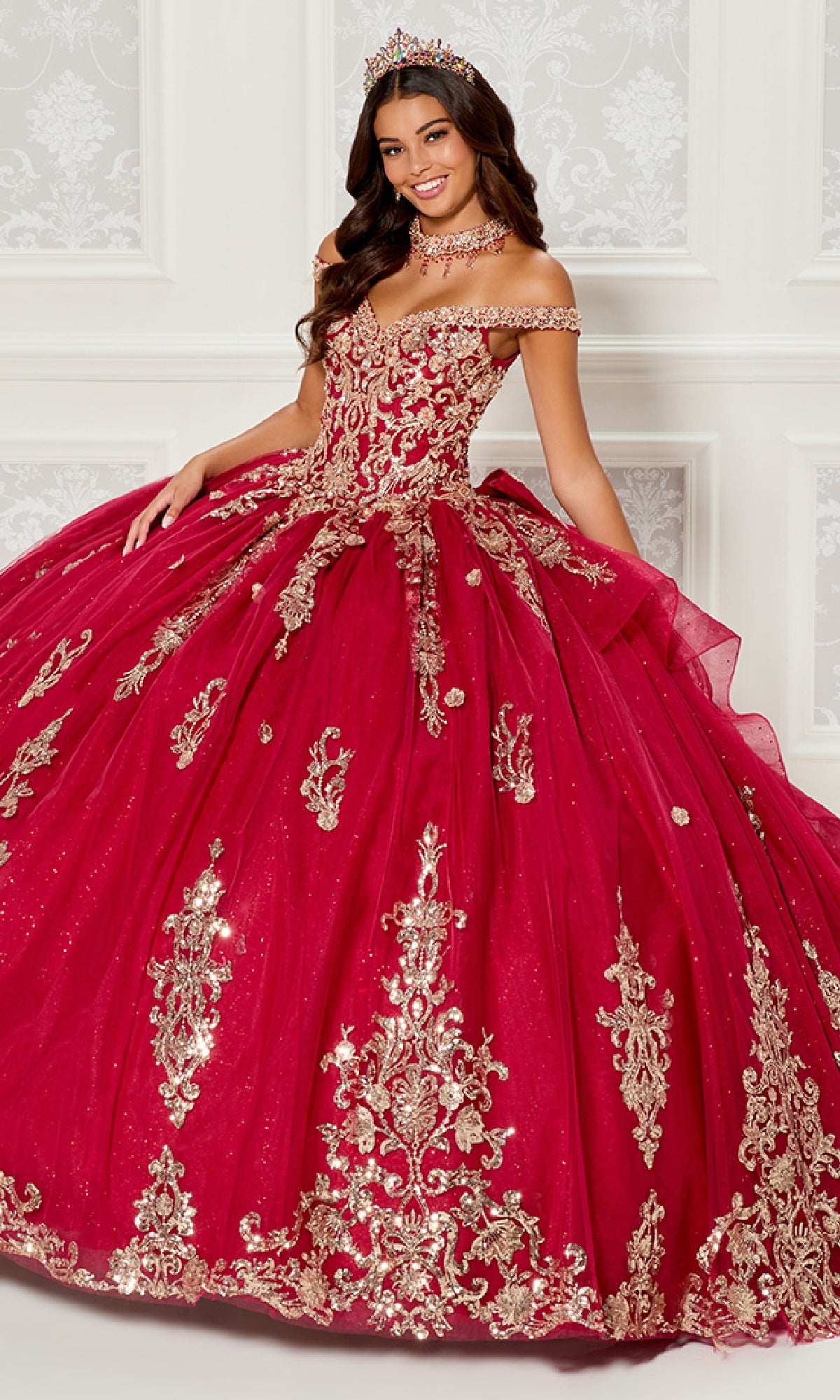 A woman in the Quinceanera Dress PR30112 By Princesa, an elaborate red gown with gold embroidery, an off-the-shoulder neckline, and a matching crown, stands in an elegant white-paneled room.